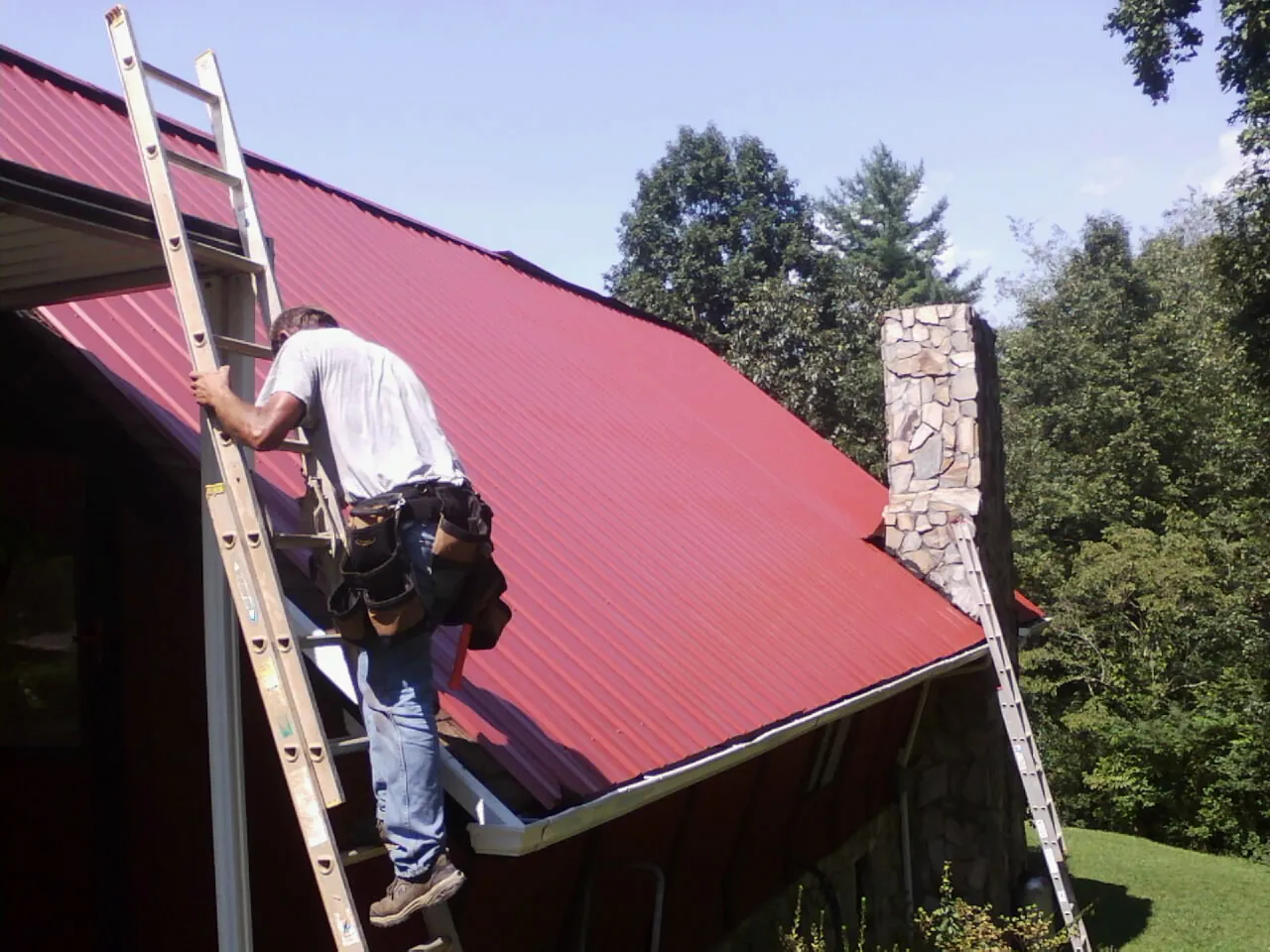 Roofer on a ladder beside a red metal roof and stone chimney — Soffit & Fascia Repair by Roofing Brothers in Dunlap